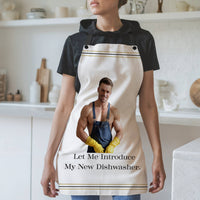 A woman wearing a white apron with black straps stands in a modern kitchen. The apron features an image of a muscular, shirtless man wearing denim overalls, yellow rubber gloves, and smiling. Below the image, the text reads: "Let Me Introduce My New Dishwasher." The apron has thin black and gold stripes along the top and bottom edges. The background includes white shelves, cutting boards, and kitchen items.