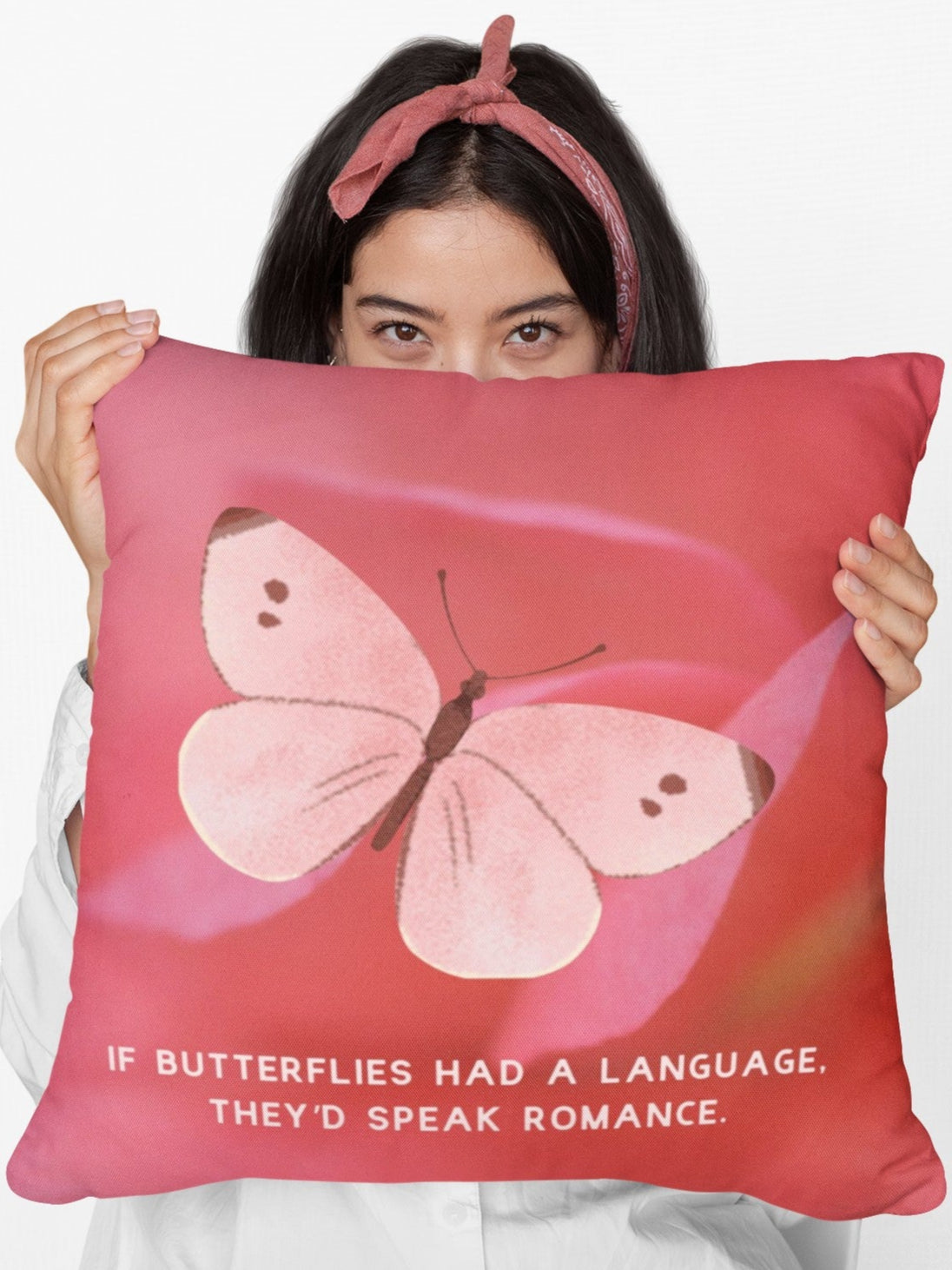 A woman holding a 20 inch pillow featuring a soft pink butterfly with delicate brown markings on its wings, set against a blurred close-up of a pink flower petal. The butterfly has a watercolor-like texture and a graceful appearance. Below the butterfly, pale pink uppercase text reads: "If butterflies had a language, they’d speak romance." The background is a warm, romantic shade of pink, creating a dreamy and whimsical aesthetic.