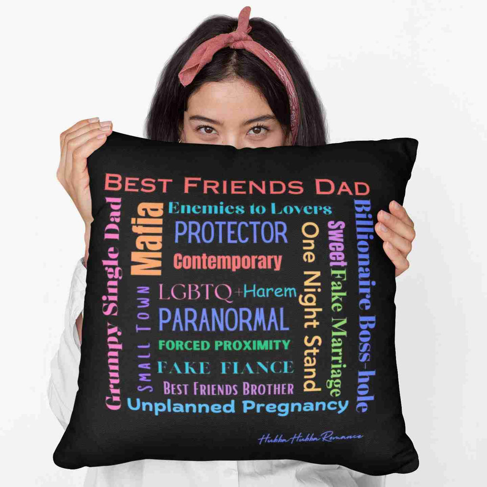 A woman holding a 20-inch square pillow features a black background with colorful text in various fonts and orientations. It lists popular romance tropes such as “Best Friends Dad,” “Enemies to Lovers,” “Grumpy Single Dad,” “Paranormal,” “Billionaire Boss-hole,” and many others, in colors like red, blue, purple, orange, pink, and green. The design forms a centered word cloud. The pillow’s backside is the same. It is soft, plush, and designed for romance readers who know their tropes—and live for them.