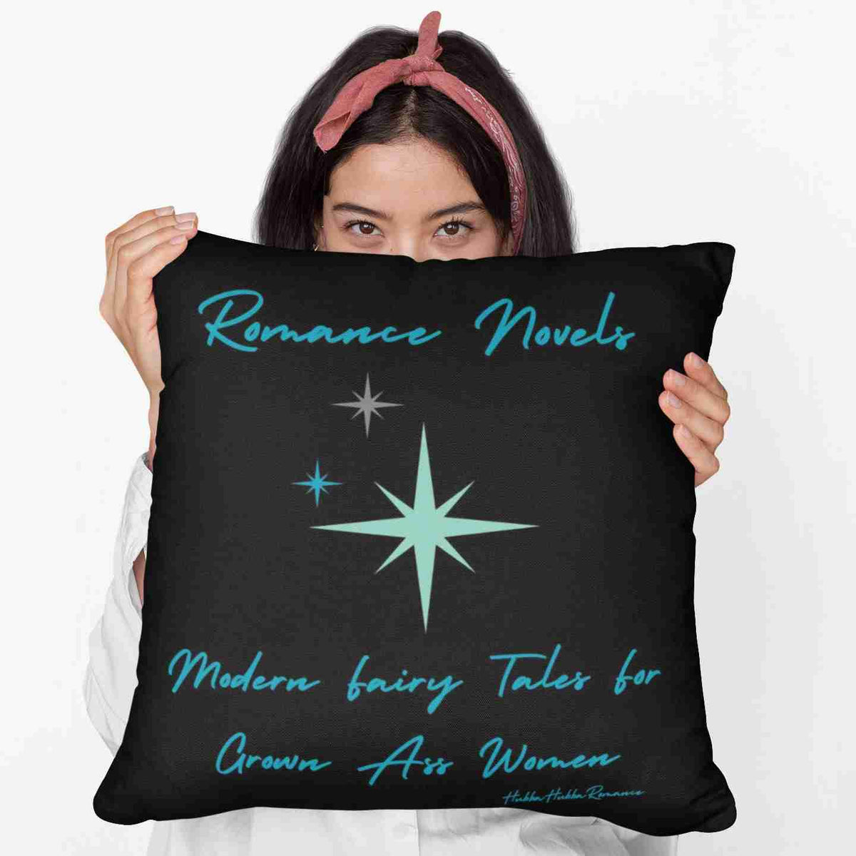 A woman holding up a 20-inch pillow featuring a bold black background with elegant teal script at the top reading: "Romance Novels." Below, in a playful, modern font, the phrase "modern fairy tales for grown-ass women" is displayed. To the left of the text, there are three teal starbursts in varying sizes, adding a vintage-inspired, whimsical touch. The minimalist yet striking design conveys a love for romance books with a sassy, empowering attitude.

