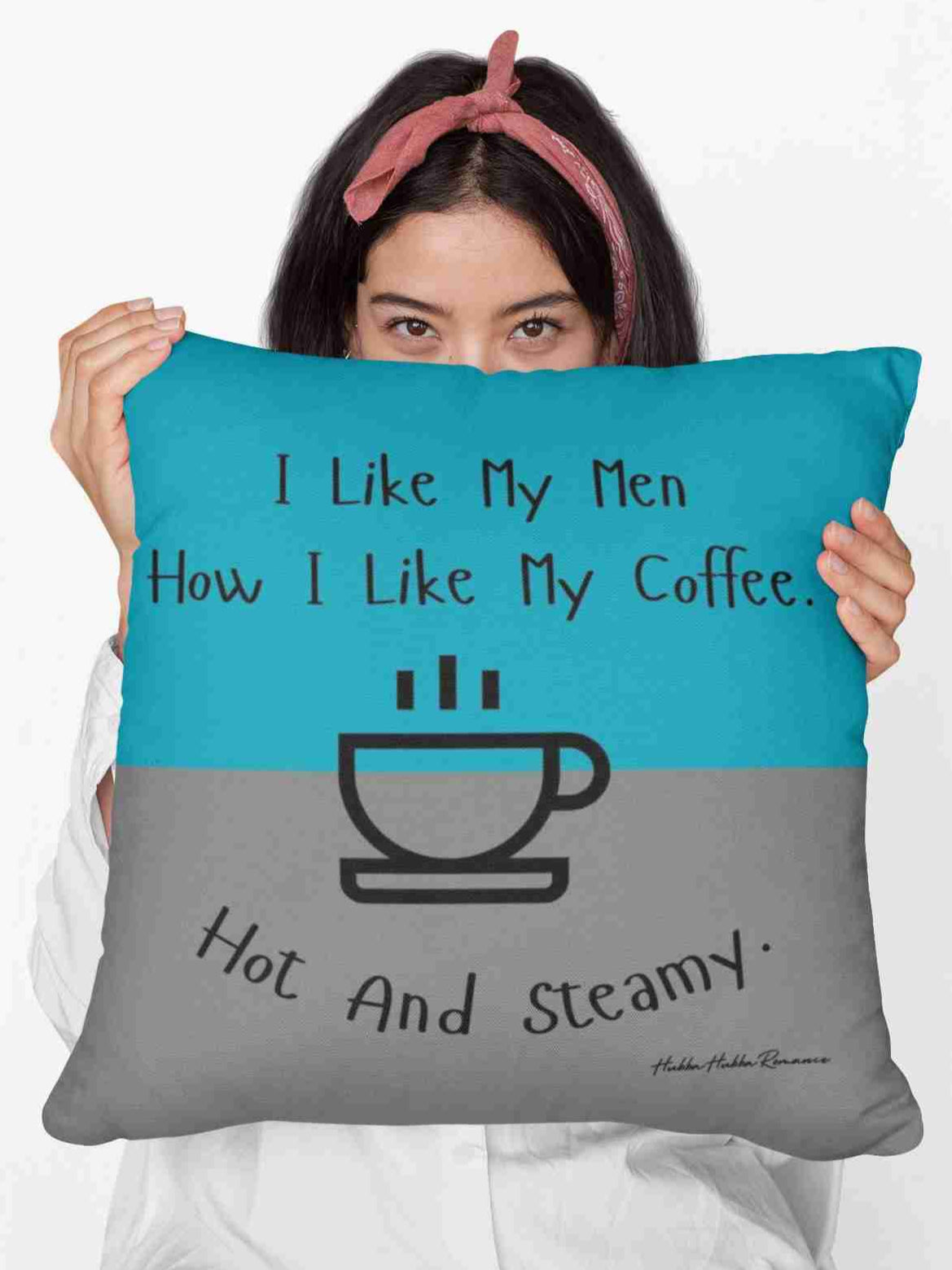 A woman holding a 20” pillow that features a split background with the top half in teal and the bottom half in gray. Centered at the top, black handwritten-style text reads: "I like my men how I like my coffee." Below, there is a simple black line drawing of a steaming coffee cup. Beneath the cup, curved text in black reads: "Hot and steamy." The design is minimalistic and playful, with a flirty and humorous tone. 