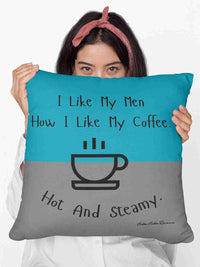 A woman holding a 20” pillow that features a split background with the top half in teal and the bottom half in gray. Centered at the top, black handwritten-style text reads: "I like my men how I like my coffee." Below, there is a simple black line drawing of a steaming coffee cup. Beneath the cup, curved text in black reads: "Hot and steamy." The design is minimalistic and playful, with a flirty and humorous tone. 