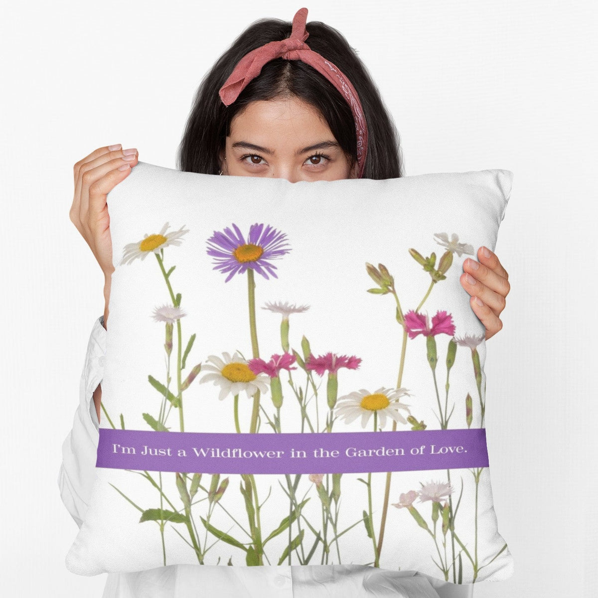 A woman is holding a 20-inch pillow that features a collection of wildflowers, including daisies, pink carnations, and a prominent purple aster, set against a clean white background. A horizontal purple banner stretches across the center, displaying the phrase "I'm Just a Wildflower in the Garden of Love." in white text. The design has a soft, romantic feel, evoking themes of individuality, natural beauty, and love.