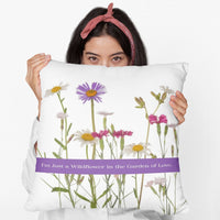 A woman is holding a 20-inch pillow that features a collection of wildflowers, including daisies, pink carnations, and a prominent purple aster, set against a clean white background. A horizontal purple banner stretches across the center, displaying the phrase "I'm Just a Wildflower in the Garden of Love." in white text. The design has a soft, romantic feel, evoking themes of individuality, natural beauty, and love.