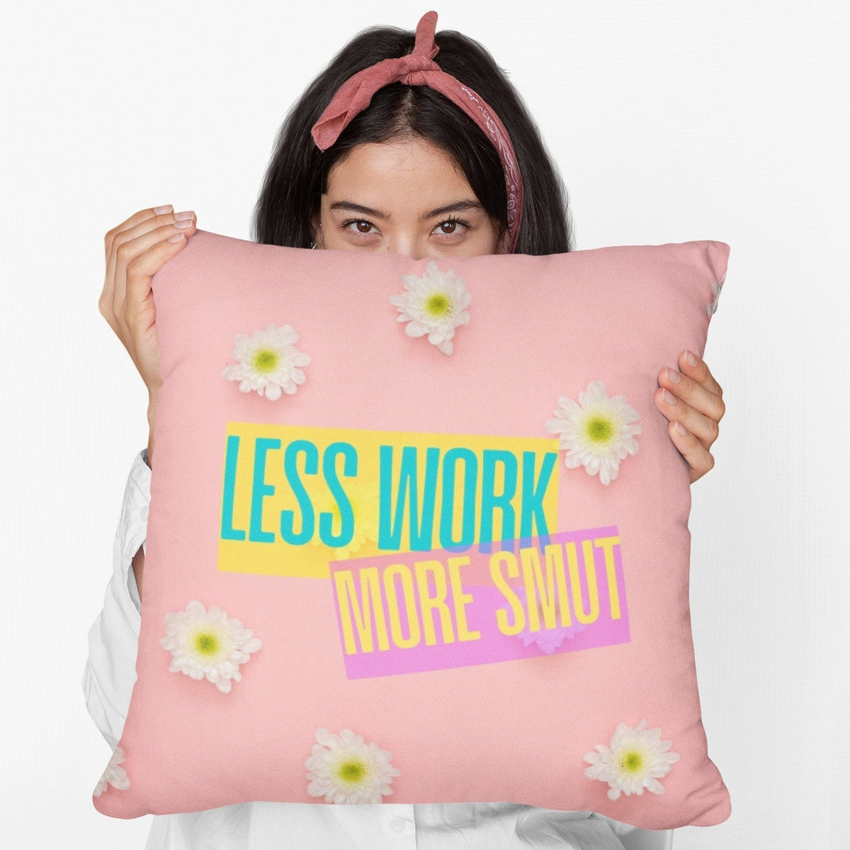 A woman holds a 20-inch pillow featuring a pastel pink background scattered with fresh white daisies. Bold, colorful text overlays the center, reading "LESS WORK" in bright teal on a yellow background and "MORE SMUT" in hot pink on a purple background. The playful, modern typography and vibrant color contrast give the design a fun, sassy, and energetic vibe, perfect for romance readers who prioritize their book boyfriends over their to-do lists.