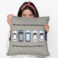 An image of a female holding a 20-inch pillow featuring an overhead view of a row of parked cars in a parking lot. Above the image, bold black text reads: "Men Are Like Parking Spaces." Below the image, matching text continues: "All The Good Ones Are Taken." The background is a muted grayish-olive greenish, allowing the text and image to stand out. The design is playful and sarcastic, making a humorous comparison between dating and the struggle to find an available parking spot. 