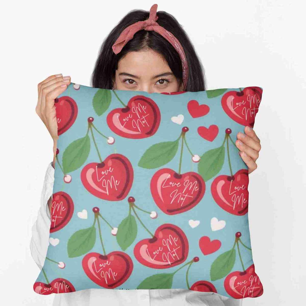 A woman is holding a 20-inch pillow that features a playful pattern of heart-shaped cherries with green leaves and stems against a light blue background. Scattered among the cherries are small red and pink hearts. Each cherry is adorned with elegant white cursive text that alternates between "Love Me" and "Love Me Not," evoking the classic game of romantic uncertainty. The whimsical, romantic design combines a sweet aesthetic with a touch of flirtatious fun.
