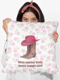 This square pillow features a pair of brown leather cowboy boots with intricate stitching and metal accents, topped with a red cowboy hat with black stitched detailing. The background is soft white with a delicate pink floral pattern. Below the boots and hat, the bold brown text reads: "When cowboy boots meets cowgirl sass!" The design combines Western charm with a playful, feminine flair.