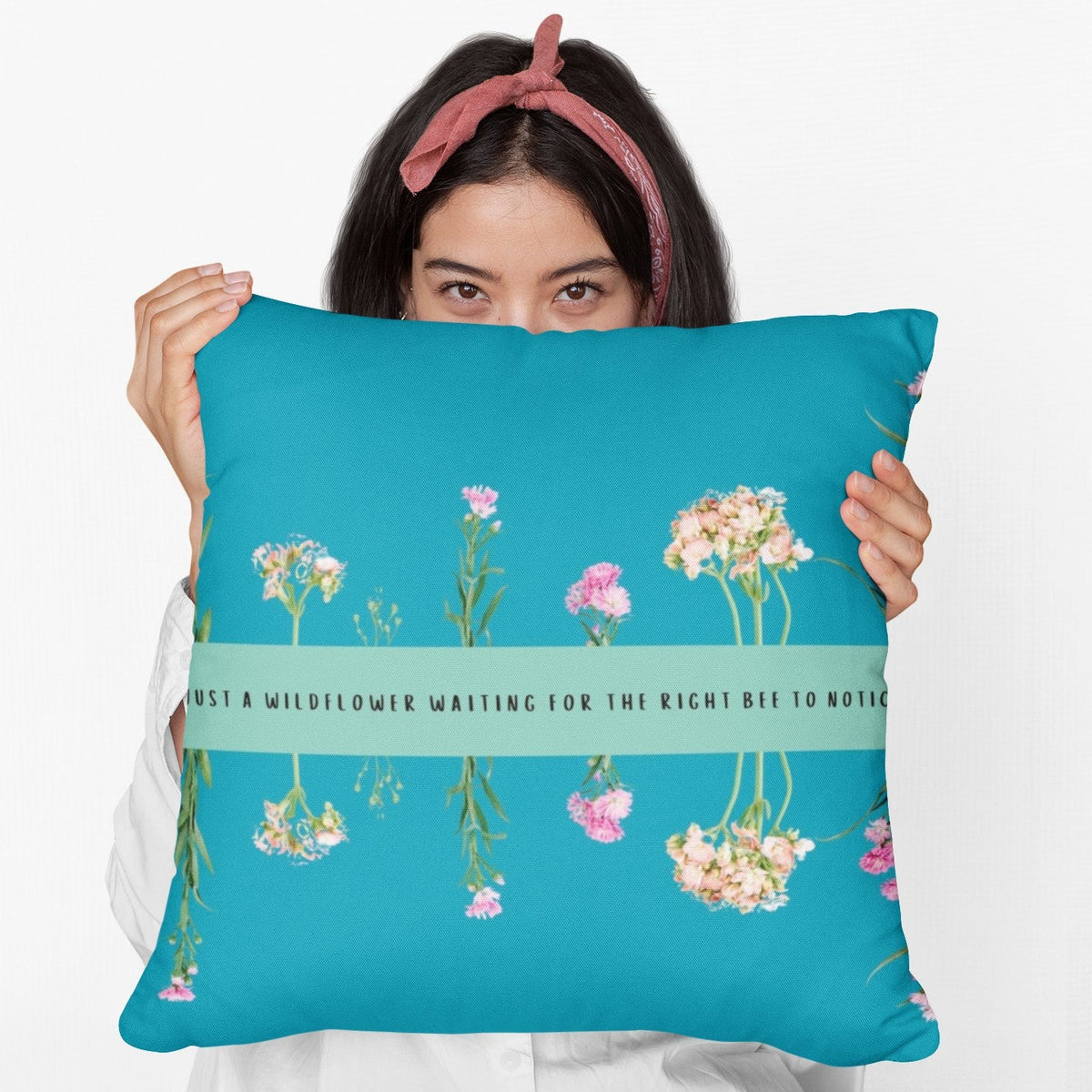 A woman holds a 20- inch pillow that features an arrangement of delicate wildflowers, some upright and others hanging upside down, against a vibrant teal background. A horizontal light green banner stretches across the center, displaying the phrase "I'm just a wildflower waiting for the right bee to notice." in black text. The design is whimsical and romantic, symbolizing patience, individuality, and the hope of finding the perfect match.