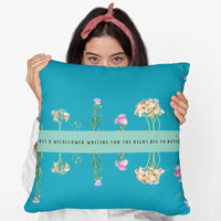 A woman holds a 20- inch pillow that features an arrangement of delicate wildflowers, some upright and others hanging upside down, against a vibrant teal background. A horizontal light green banner stretches across the center, displaying the phrase "I'm just a wildflower waiting for the right bee to notice." in black text. The design is whimsical and romantic, symbolizing patience, individuality, and the hope of finding the perfect match.