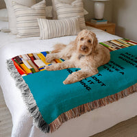 The blanket is displayed neatly folded on a bed with a white bedspread and striped pillows and a large white dog laying on it.
