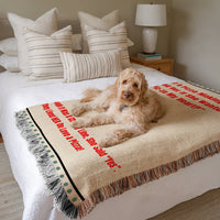 The blanket is displayed neatly folded on a bed with a white bedspread and striped pillows and a large white dog laying on it.
