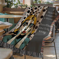 The blanket is displayed lying on a chair in a coffee shop setting with an open book and a cup of coffee on a small round table.
