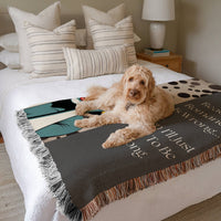 The blanket is displayed neatly folded on a bed with a white bedspread and striped pillows and a large white dog laying on it.
