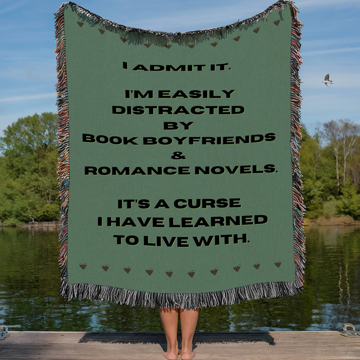 This cozy woven blanket features a playful quote about being easily distracted by book boyfriends and romance novels, surrounded by small heart icons at the top and bottom of the blanket on a muted green background with bold black text. The edges are trimmed in colorful cotton fringe. It is being displayed by a woman who is showing it outstretched behind her. She is standing on a lakefront dock in a lake setting. The only visible part of her are her legs.   