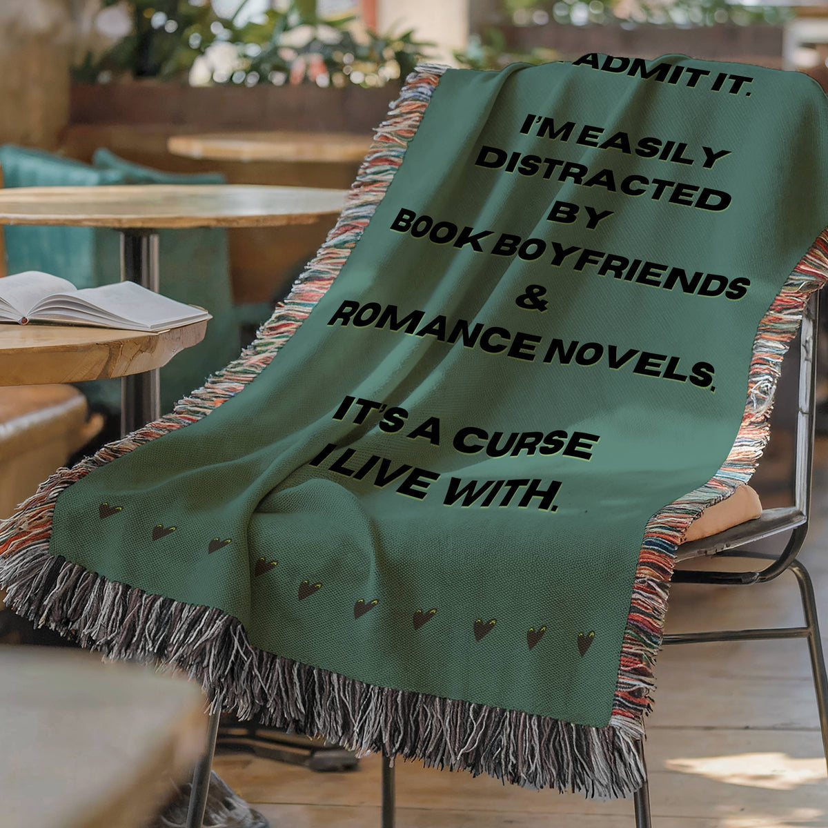 The blanket is displayed lying on a chair in a coffee shop setting with an open book and a cup of coffee on a small round table.

