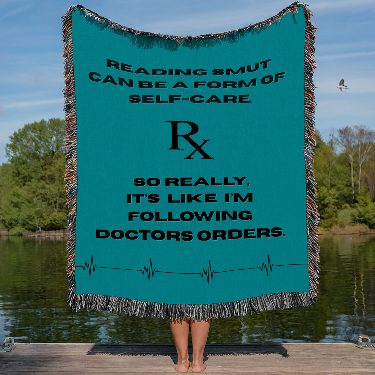 This woven blanket, being modeled by someone standing on a lake dock, holding the blanket stretched out behind them, features a soft blue background with a playful quote in bold black text: “Reading Smut Can Be A Form Of Self-Care.” Centered underneath is a large RX medical symbol. Underneath the Rx symbol is the rest of the quote *So Really I’m Just Following Doctor’s Orders.” At the bottom of the blanket is a “heart line” graphic mimicking someone's heart rate. It has a colorful fringed edge all around.