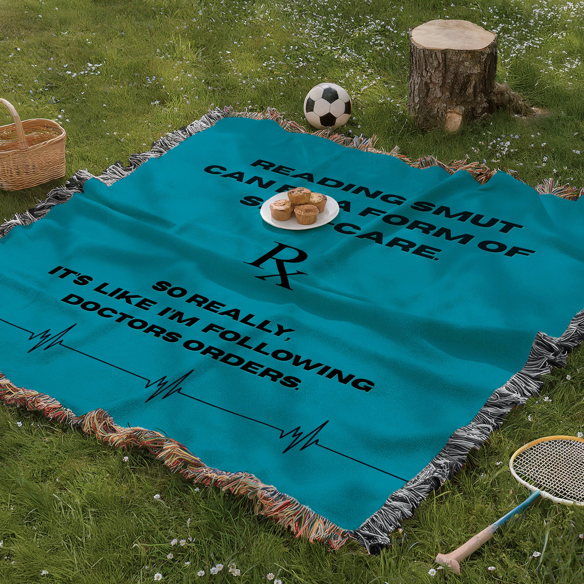 The blanket is being displayed as a picnic blanket in a park setting with a tree stump, picnic basket, plate of muffins, and a tennis racquet and a soccer ball lying around.

