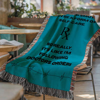 The blanket is displayed lying on a chair in a coffee shop setting with an open book and a cup of coffee on a small round table.


