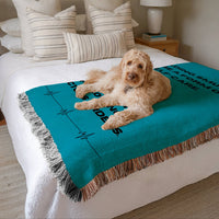 The blanket is displayed neatly folded on a bed with a white bedspread and striped pillows and a large white dog lying on it.

