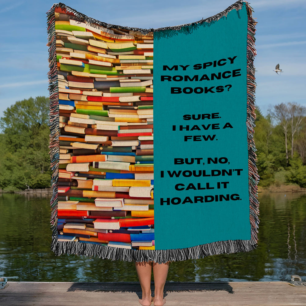 This woven blanket features a colorful, vertical, chaotic stack of books on the right side and a turquoise background on the left side ,with the following phrase stacked vertically, “My spicy romance books? Sure. I have a few. But I wouldn’t call it hoarding” . The blanket is displayed by a woman who is showing it outstretched behind her. She is standing on a lakefront dock in a lake setting with only  her legs showing.