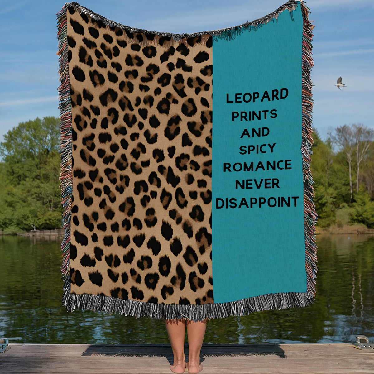 This cozy woven blanket features a bold black, tan and brown leopard print pattern on one half while the other half has a turquoise blue background with the phrase “Leopard Prints and Spicy Romance Never Disappoint” in a bold black font. It is being displayed by a woman who is showing it outstretched behind her. She is standing on a lakefront dock in a lake setting. The only visible part of her are her legs.  