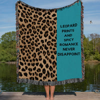 This cozy woven blanket features a bold black, tan and brown leopard print pattern on one half while the other half has a turquoise blue background with the phrase “Leopard Prints and Spicy Romance Never Disappoint” in a bold black font. It is being displayed by a woman who is showing it outstretched behind her. She is standing on a lakefront dock in a lake setting. The only visible part of her are her legs.  