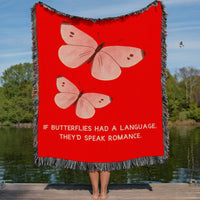 This cozy woven blanket features two charming, beautiful pink butterflies on a reddish orange background with text in a soft pink that asks “If butterflies had a love language, they’d speak romance.” The blanket has colorful cotton fringe around the edges. It is being displayed by a woman who is showing it outstretched behind her. She is standing on a lakefront dock in a lake setting. The only visible part of her are her legs.   