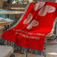 The blanket is displayed lying on a chair in a coffee shop setting with an open book and a cup of coffee on a small round table.
