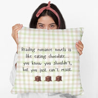 A woman is holding the square pillow with a beige , pink and green plaid background. Black playful text says: “Reading romance novels is like eating chocolate… you know you shouldn’t, but you just can’t resist.” Three illustrated chocolate pieces sit along the bottom. A cheeky decorative pillow for romance readers.