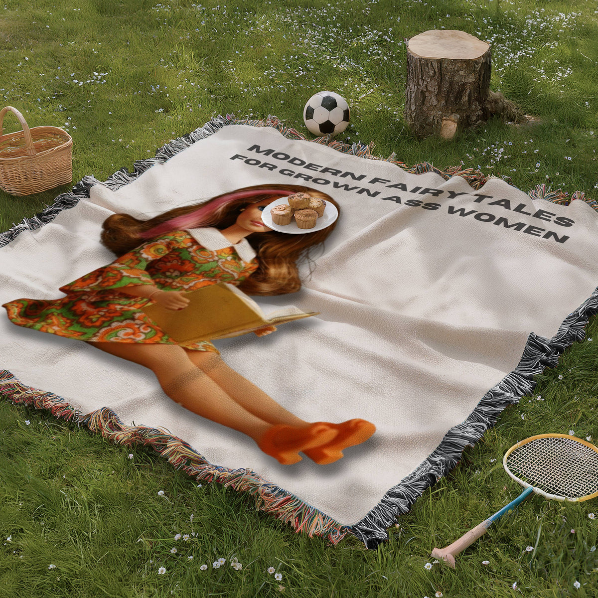 Vintage-inspired woven blanket spread on green grass featuring a retro brunette reader doll with a pink streak in her hair and the phrase “Modern Fairy Tales for Grown Ass Women.” Cozy romance reader blanket displayed in an outdoor picnic setting with basket, tree stump, and badminton racket.