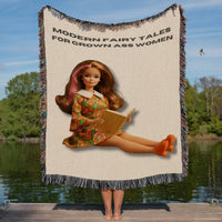 Woven cotton fringe blanket featuring a retro book-loving heroine reading a novel beneath the phrase “Modern Fairy Tales for Grown Ass Women.” Blanket displayed outdoors against a lake backdrop, showing the full design and fringe details for romance readers.