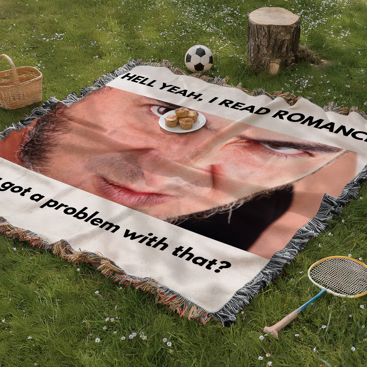 Novelty woven blanket spread on grass featuring a large dramatic male face and statement he reads romance novels and so what.