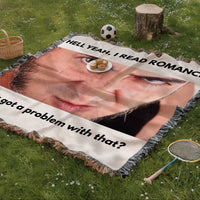 Novelty woven blanket spread on grass featuring a large dramatic male face and statement he reads romance novels and so what.