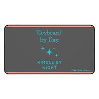 A charcoal gray desk mat features a playful message split across two lines: "Keyboard by Day" in turquoise serif text on the left, and "Kindle by Night" in sans-serif turquoise on the right. Turquoise starburst-style sparkles accent the design, and two thin orange horizontal lines run along the top and bottom edges. This is the medium version.