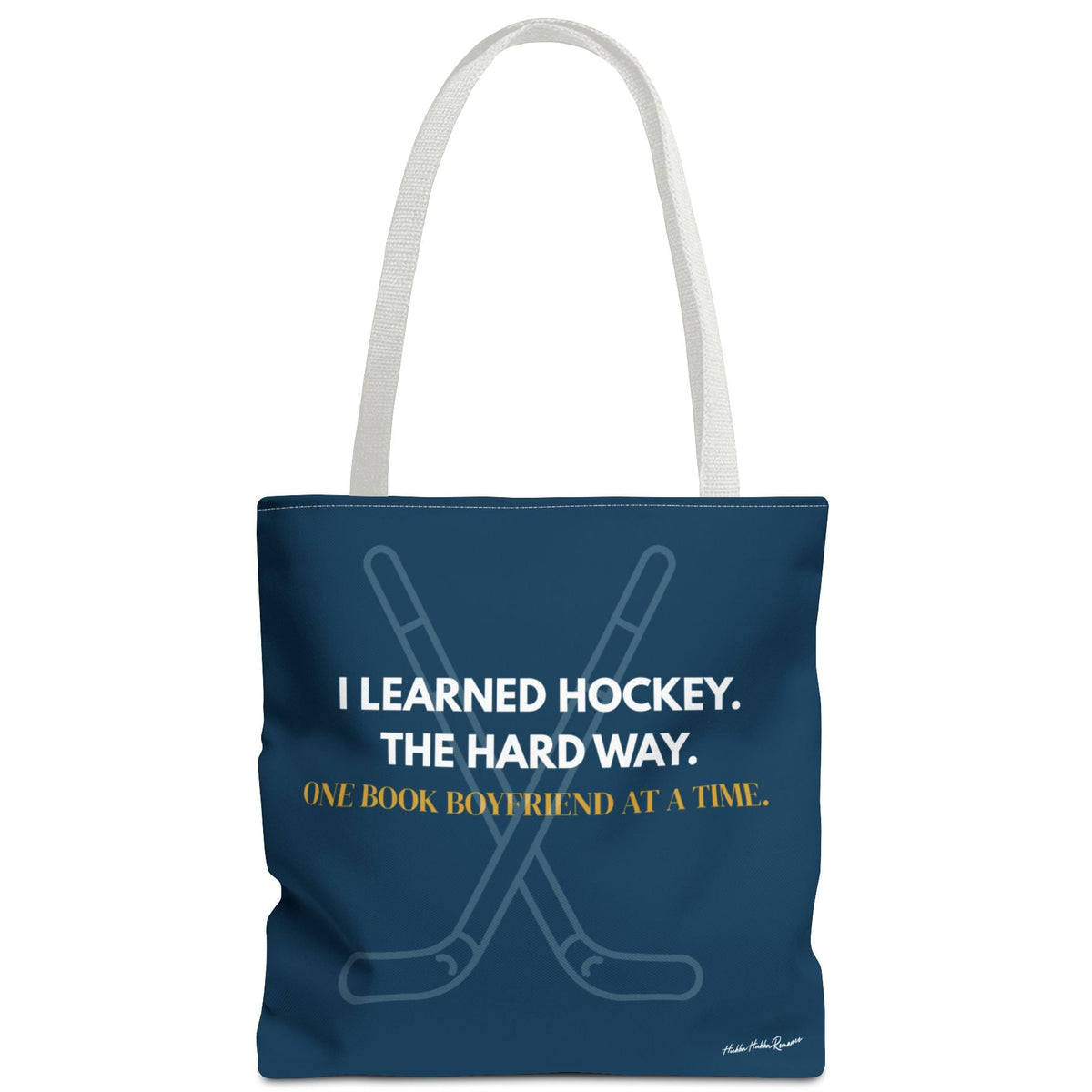 Navy blue hockey romance tote bag with white handles, crossed hockey sticks and the words “I Learned Hockey. The Hard Way” in white and "One Book Boyfriend at a Time" in gold.