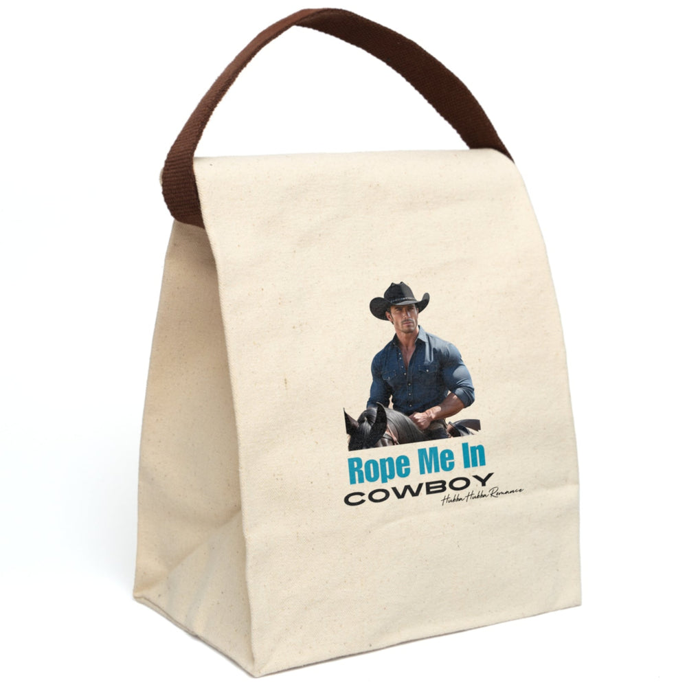 This canvas lunch bag features a full-color image of a muscular, chiseled cowboy riding a black horse. He wears a fitted denim button-down shirt and a black cowboy hat with silver details. His arms are crossed on the saddle, and he gazes ahead with intensity. Below the image, bold turquoise text reads “Rope Me In” followed by black block letters that say “COWBOY.” The bag includes a canvas strap and roll-top closure. The strap is displyed upright.