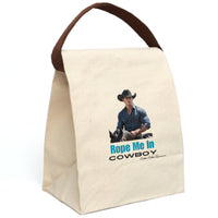 This canvas lunch bag features a full-color image of a muscular, chiseled cowboy riding a black horse. He wears a fitted denim button-down shirt and a black cowboy hat with silver details. His arms are crossed on the saddle, and he gazes ahead with intensity. Below the image, bold turquoise text reads “Rope Me In” followed by black block letters that say “COWBOY.” The bag includes a canvas strap and roll-top closure. The strap is displyed upright.