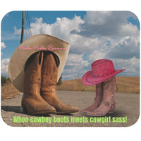 This mouse pad features two pairs of cowboy boots facing each other. One pair is for a guy and is slightly dusty. A large tan cowboy hat is hanging over the top of the boots. The second pair is clearly for a female, and there is a pink cowboy hat resting on top of the boots. Both pairs are sitting on an old country road with blue skies in the background.