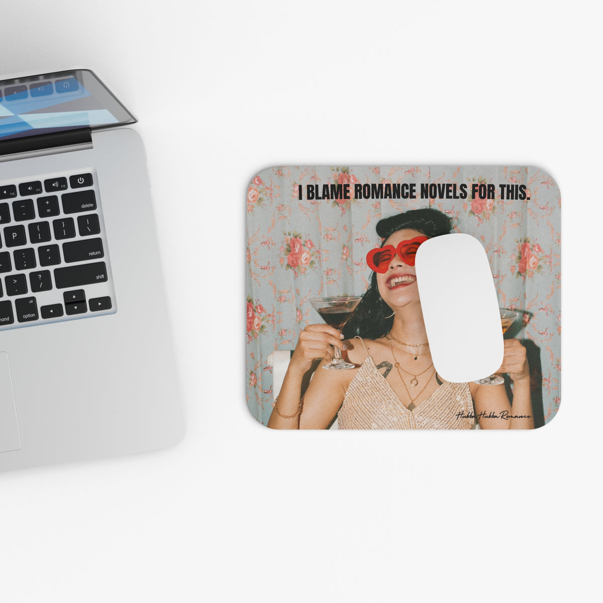 A rectangular mouse pad with a retro-style illustration f a woman and the text 'I BLAME ROMANCE NOVELS FOR THIS.' shown on a white desk next to a laptop.