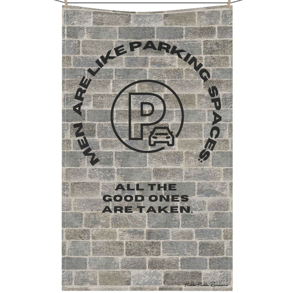 The image depicts a cotton kitchen towel hanging on a clothesline with a vintage-style design, set against a textured brick wall background. At the top, arched black text reads: "Men Are Like Parking Spaces." Below, a circular stamp-like emblem resembles a classic parking permit, with a distressed, worn effect. The stamp has bold block letters and a faded appearance, enhancing its retro feel. Beneath the stamp, additional black text reads: "All the Good Ones Are Taken." The combination of the brick texture 