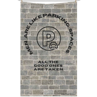 The image depicts a cotton kitchen towel hanging on a clothesline with a vintage-style design, set against a textured brick wall background. At the top, arched black text reads: "Men Are Like Parking Spaces." Below, a circular stamp-like emblem resembles a classic parking permit, with a distressed, worn effect. The stamp has bold block letters and a faded appearance, enhancing its retro feel. Beneath the stamp, additional black text reads: "All the Good Ones Are Taken." The combination of the brick texture 