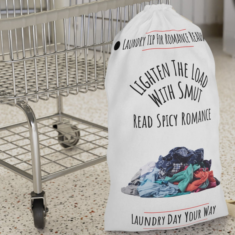 This laundry bag features a white background with a colorful pile of clothes at the bottom. Above the pile, black text reads, “Lighten the Load with Smut” in an arched layout. Below that, in a straight line, it says: “Read Spicy Romance.” At the top, the header “Laundry Tip for Romance Readers” is centered between two black dots and underlined in red. The bottom of the image reads: “Laundry Day Your Way” in black text, also underlined in red. The bag includes a drawstring closure and a canvas carry strap.