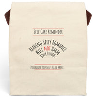 This canvas lunch bag design features black and red text on a white background. At the top, bold lettering reads: “Self-Care Reminder,” underscored with a red line. The main message is displayed in a curved arch and reads, “Reading Spicy Romance Will Not Harm Your Lunch,” with the word “Not” highlighted in red. At the bottom, text reads: “Prioritize Yourself. Read More,” also framed with red lines. The bag has a roll-top closure and a sturdy canvas carrying strap.