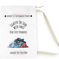 This laundry bag features a white background with a colorful pile of clothes at the bottom. Above the pile, black text reads: “Lighten the Load with Smut” in an arched layout. Below that, in a straight line, it says: “Read Spicy Romance.” At the top, the header “Laundry Tip for Romance Readers” is centered between two black dots and underlined in red. The bottom of the image reads: “Laundry Day Your Way” in black text, also underlined in red. The bag includes a drawstring closure and a canvas carry strap.
