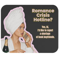 A rectangular mouse pad with rounded corners featuring a dark gray background, a cutout photo of a woman in a white towel wrap holding a phone and a champagne bottle, and the text 'Romance Crisis Hotline? Yes, hi, I’d like to report a shortage of book boyfriends.' in pink lettering with a gold outline.