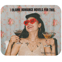 A rectangular mouse pad with rounded corners featuring a vintage-style illustration of a woman holding two cocktails, wearing red heart-shaped sunglasses, with the text 'I BLAME ROMANCE NOVELS FOR THIS.' at the top.