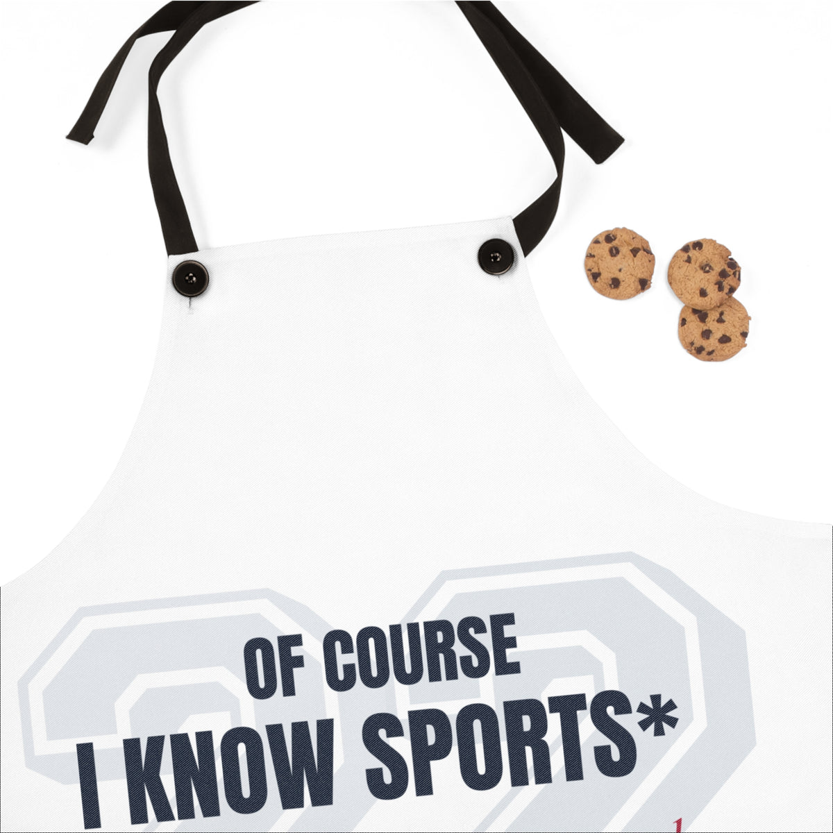 White novelty apron with black neck strap and two black buttons, featuring the text 'OF COURSE I KNOW SPORTS*' in dark navy over a light gray athletic number graphic. Three chocolate chip cookies are placed to the right as props.