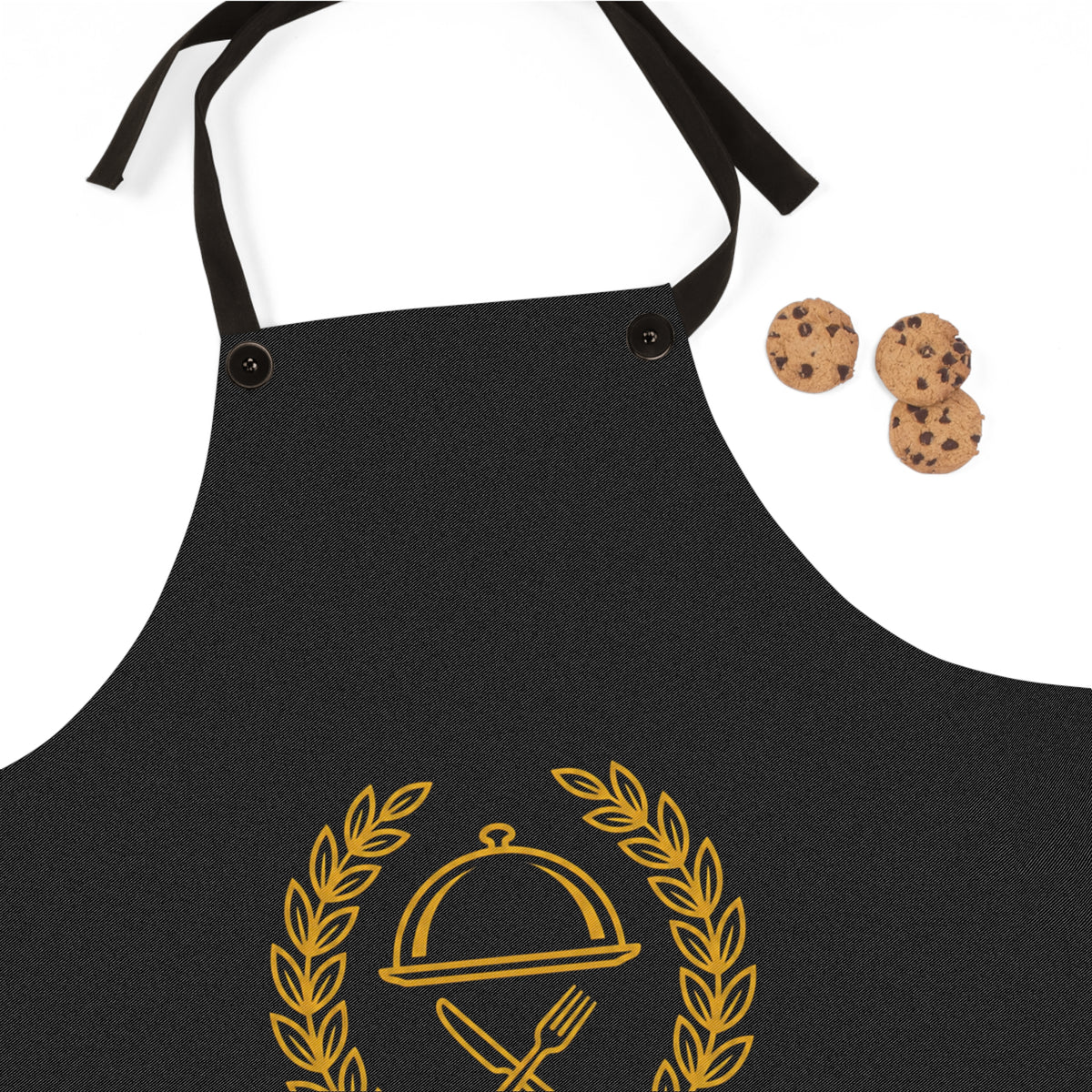 A black kitchen apron with a gold-colored cloche and fork graphic, shown on a white background with three chocolate chip cookies as props.