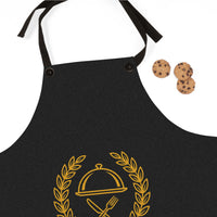 A black kitchen apron with a gold-colored cloche and fork graphic, shown on a white background with three chocolate chip cookies as props.